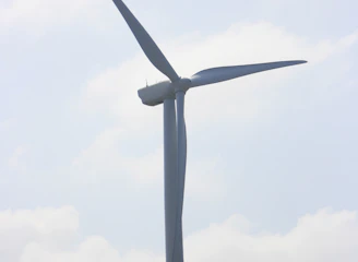 A large wind turbine stands prominently against a partly cloudy sky. It towers above grassy hills, indicating a renewable energy source in a natural landscape.
