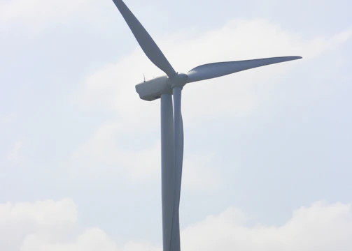 A large wind turbine stands prominently against a partly cloudy sky. It towers above grassy hills, indicating a renewable energy source in a natural landscape.