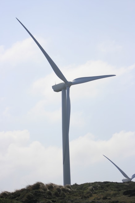 A large wind turbine stands prominently against a partly cloudy sky. It towers above grassy hills, indicating a renewable energy source in a natural landscape.