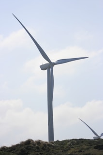 A large wind turbine stands prominently against a partly cloudy sky. It towers above grassy hills, indicating a renewable energy source in a natural landscape.