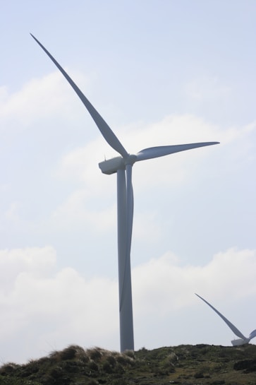 A large wind turbine stands prominently against a partly cloudy sky. It towers above grassy hills, indicating a renewable energy source in a natural landscape.