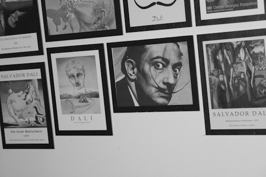 Black and white photograph of a wall featuring framed images and artworks, primarily associated with Salvador Dal&iacute;. Most images are surrealist paintings and drawings, including a prominent portrait of a man with exaggerated facial features and a mustache.