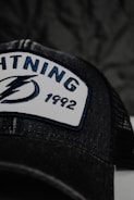 A close-up of a black cap featuring an embroidered patch with the word 'LIGHTNING', a stylized lightning bolt logo, and the number '1992'. The patch has a white background with blue and black embroidery. The cap has mesh detailing visible in the image.