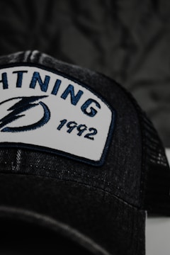 A close-up of a black cap featuring an embroidered patch with the word 'LIGHTNING', a stylized lightning bolt logo, and the number '1992'. The patch has a white background with blue and black embroidery. The cap has mesh detailing visible in the image.