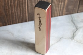 A rectangular block made of wood with the brand name 'schlagwerk' engraved on it. The sides feature layers of colored wood, with visible red and brown tones, resting on a gray marble-like surface.