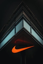 A modern, minimalist architectural structure with reflective glass panels meeting at a sharp corner. An illuminated orange swoosh logo is prominently displayed on the building's facade, contrasting against the dark surroundings.