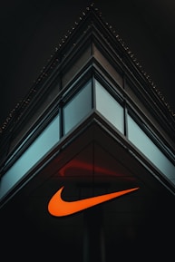 A modern, minimalist architectural structure with reflective glass panels meeting at a sharp corner. An illuminated orange swoosh logo is prominently displayed on the building's facade, contrasting against the dark surroundings.