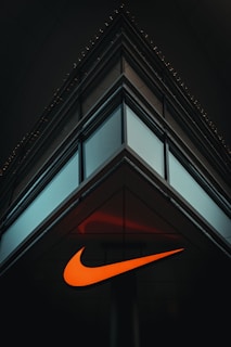 A modern, minimalist architectural structure with reflective glass panels meeting at a sharp corner. An illuminated orange swoosh logo is prominently displayed on the building's facade, contrasting against the dark surroundings.