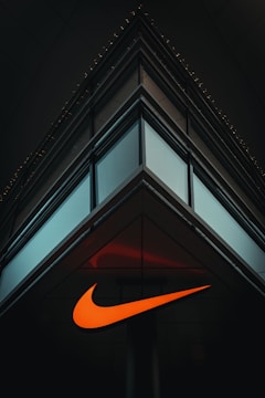 A modern, minimalist architectural structure with reflective glass panels meeting at a sharp corner. An illuminated orange swoosh logo is prominently displayed on the building's facade, contrasting against the dark surroundings.