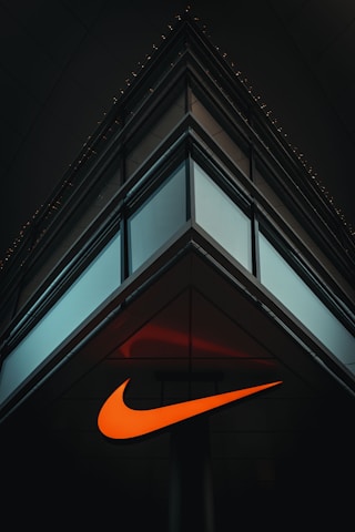 A modern, minimalist architectural structure with reflective glass panels meeting at a sharp corner. An illuminated orange swoosh logo is prominently displayed on the building's facade, contrasting against the dark surroundings.