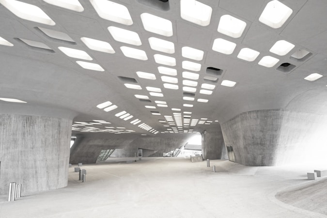 A modern, spacious architectural interior with large, curved concrete surfaces. The ceiling features a grid of rectangular light panels, casting bright, diffused light onto the otherwise subdued gray structure. The space appears open with minimal furnishings and a futuristic aesthetic.