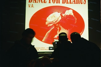 A poster with the text 'Dance for Belarus V.A.' features a red and white design with an image of a hand holding a rose. Three silhouetted figures appear in the foreground, facing the poster.