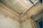 A detailed view of a deteriorating room corner featuring ornate ceiling moldings with floral designs, patches of peeling paint on the walls, and visible structural damage. A modern metallic pipe with two round ventilation outlets runs along the ceiling, coupled with an industrial-style light fixture.