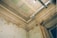 A detailed view of a deteriorating room corner featuring ornate ceiling moldings with floral designs, patches of peeling paint on the walls, and visible structural damage. A modern metallic pipe with two round ventilation outlets runs along the ceiling, coupled with an industrial-style light fixture.