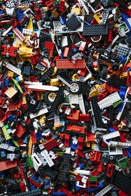 A chaotic and colorful assortment of interlocking plastic building toy bricks, shown in a variety of sizes and shapes, scattered randomly. The pieces are predominantly made up of bright reds, blues, yellows, whites, and blacks.