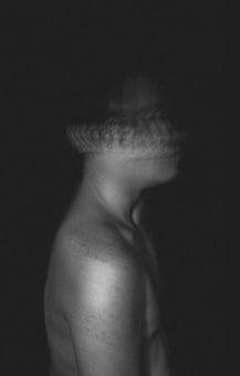 A blurred grayscale image of a shirtless person with their head appearing in motion, suggesting sideways movement. The image is dark, with little detail visible beyond the contours of the body.