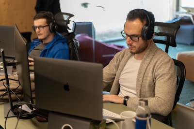 Two people are sitting at desks in an office environment, each working on a laptop. They are both wearing headphones and appear to be focused on their tasks. The workspace has a modern feel with ergonomic chairs and large monitors.