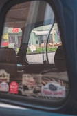 The image captures the interior of a car through a partially open window, reflecting a street scene outside. Various colorful stickers are displayed on the window. The background includes a building with green tones and outdoor seating arrangements, visible through the window's reflection.