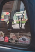 The image captures the interior of a car through a partially open window, reflecting a street scene outside. Various colorful stickers are displayed on the window. The background includes a building with green tones and outdoor seating arrangements, visible through the window's reflection.