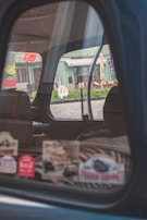 The image captures the interior of a car through a partially open window, reflecting a street scene outside. Various colorful stickers are displayed on the window. The background includes a building with green tones and outdoor seating arrangements, visible through the window's reflection.