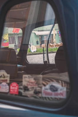 The image captures the interior of a car through a partially open window, reflecting a street scene outside. Various colorful stickers are displayed on the window. The background includes a building with green tones and outdoor seating arrangements, visible through the window's reflection.