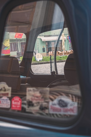 The image captures the interior of a car through a partially open window, reflecting a street scene outside. Various colorful stickers are displayed on the window. The background includes a building with green tones and outdoor seating arrangements, visible through the window's reflection.