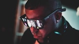 A person is wearing large, futuristic sunglasses with LED lights that display patterns on the lenses. They are also wearing headphones around their neck. The background is dark, enhancing the glow from the glasses.