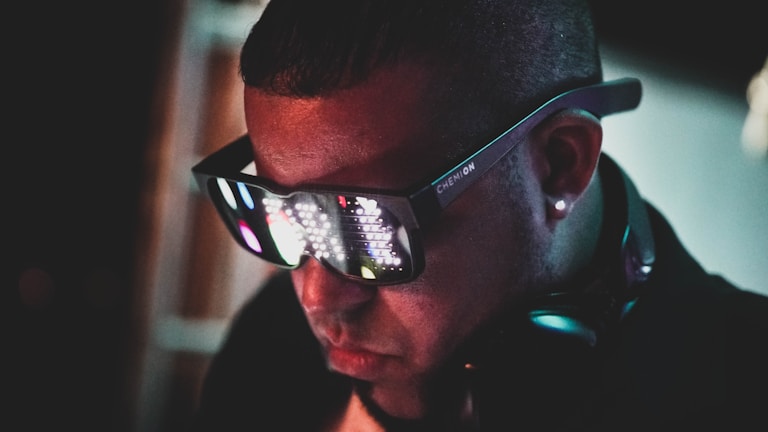 A person is wearing large, futuristic sunglasses with LED lights that display patterns on the lenses. They are also wearing headphones around their neck. The background is dark, enhancing the glow from the glasses.