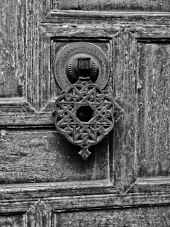 An intricately designed metal door knocker featuring a geometric and floral pattern is mounted on a weathered wooden door with visible grain and texture. The design is ornate and detailed, showcasing skilled craftsmanship.