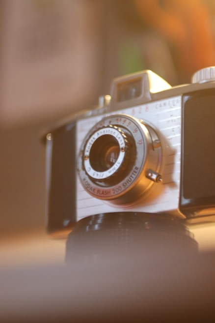 A vintage camera with a prominent lens sits in a softly lit room, capturing the nostalgia of analog photography. The camera has a classic design, featuring a metallic finish and distinct engravings on the lens.