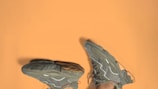 A pair of greenish-gray sneakers with intricate patterns and designs is placed against a solid light brown background. The shoes are positioned upside down, highlighting their textured soles and sleek exterior detailing.