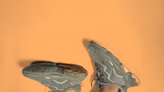 A pair of greenish-gray sneakers with intricate patterns and designs is placed against a solid light brown background. The shoes are positioned upside down, highlighting their textured soles and sleek exterior detailing.