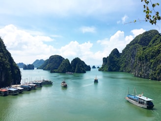 A peaceful bay featuring numerous limestone islands covered in lush greenery. Several boats are floating on the calm turquoise water, with a backdrop of a partly cloudy blue sky.