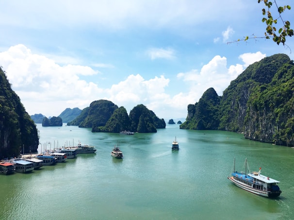 A peaceful bay featuring numerous limestone islands covered in lush greenery. Several boats are floating on the calm turquoise water, with a backdrop of a partly cloudy blue sky.