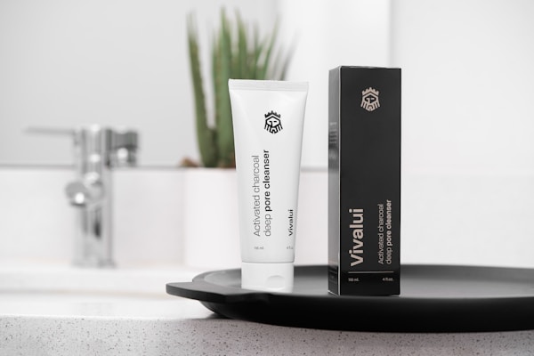 A white tube of activated charcoal deep pore cleanser labeled Vivalui stands next to its black box. Both are placed on a sleek dark tray, with a modern sink and faucet in the background, alongside a potted plant adding a natural touch.