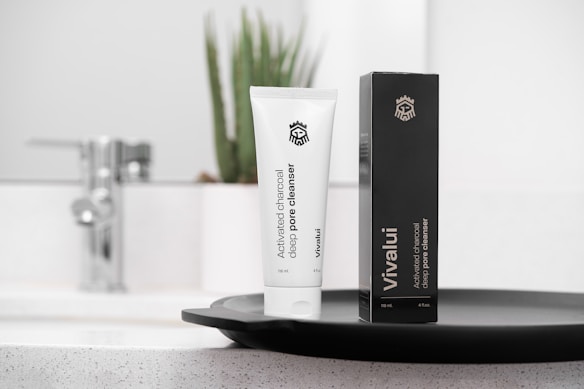 A white tube of activated charcoal deep pore cleanser labeled Vivalui stands next to its black box. Both are placed on a sleek dark tray, with a modern sink and faucet in the background, alongside a potted plant adding a natural touch.