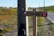 A wooden directional signpost for the 'Caminho Atlântico (via litoral)' is positioned near a concrete utility pole beside a rural road. The surrounding landscape features green and orange vegetation, and a parked white car is visible on a distant hill. The road is lined with telephone poles extending into the background.