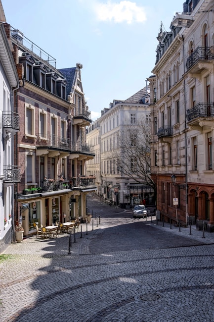 The image captures a quiet European street lined with elegant historic buildings featuring ornate balconies and classic architecture. A cobblestone road curves through the scene, leading to an intersection with parked cars and a cozy outdoor cafe with yellow chairs and tables. The buildings have large windows and are painted in muted tones of beige, cream, and light brown. Natural light casts soft shadows across the street.