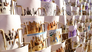 A collection of artistic prints, each depicting scenes of traditional village life, featuring desert landscapes, buildings, and people. They are arranged in an overlapping manner on display, with different color schemes and intricate designs visible.
