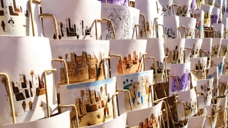 A collection of artistic prints, each depicting scenes of traditional village life, featuring desert landscapes, buildings, and people. They are arranged in an overlapping manner on display, with different color schemes and intricate designs visible.