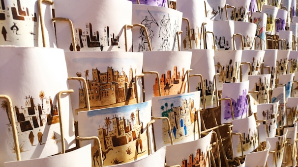 A collection of artistic prints, each depicting scenes of traditional village life, featuring desert landscapes, buildings, and people. They are arranged in an overlapping manner on display, with different color schemes and intricate designs visible.