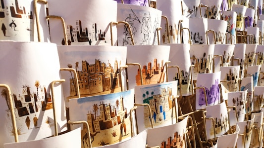 A collection of artistic prints, each depicting scenes of traditional village life, featuring desert landscapes, buildings, and people. They are arranged in an overlapping manner on display, with different color schemes and intricate designs visible.