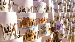 A collection of artistic prints, each depicting scenes of traditional village life, featuring desert landscapes, buildings, and people. They are arranged in an overlapping manner on display, with different color schemes and intricate designs visible.