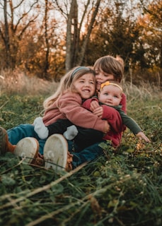 Three children are sitting together, embracing each other while smiling warmly. They are in a natural setting with grass and trees in the background, suggesting an autumn season with leaves scattered around.
