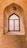 An arched, intricately designed wooden window is set in a brick wall, showcasing ornate geometric patterns. Sunlight filters through, casting detailed shadows on the surrounding bricks. The craftsmanship highlights traditional architectural styles.