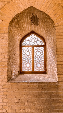 An arched, intricately designed wooden window is set in a brick wall, showcasing ornate geometric patterns. Sunlight filters through, casting detailed shadows on the surrounding bricks. The craftsmanship highlights traditional architectural styles.