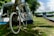 A mountain bike with large tires is leaning against a tree. Nearby, there is a blue and white tent. In the background, there's a BMX-style ramp on a grassy area, surrounded by trees and a partly cloudy sky.