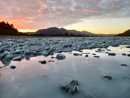 A tranquil riverbed with smooth, rounded stones scattered across the area. In the background, there's a range of hills silhouetted against a dramatic sky with colors transitioning from orange to purple, indicating either sunrise or sunset. The reflection of the colorful sky on the river adds a serene effect.