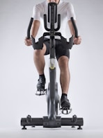 A person dressed in athletic gear is actively pedaling on a stationary exercise bike, with a focus on their lower body and cycling position. The background is minimalistic, highlighting the exercise equipment and individual.