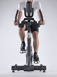 A person dressed in athletic gear is actively pedaling on a stationary exercise bike, with a focus on their lower body and cycling position. The background is minimalistic, highlighting the exercise equipment and individual.